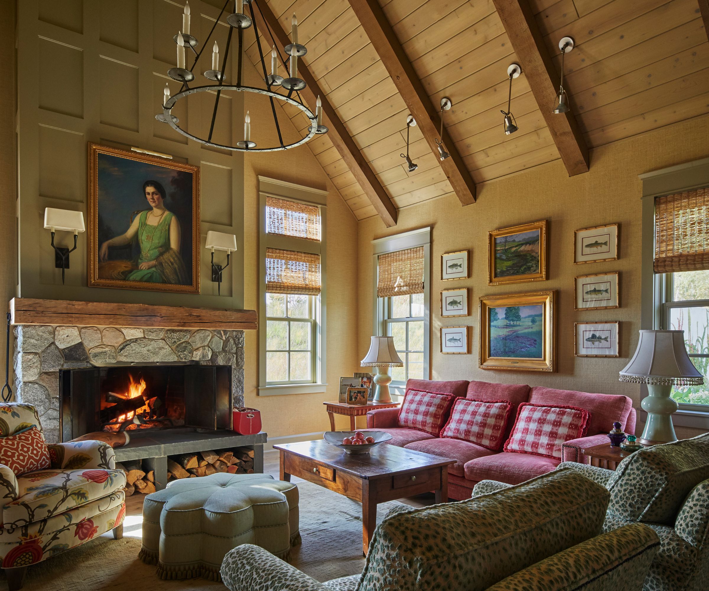 A large, cozy cabin living room with wood-clad sloped ceilings and a stone fireplace with olive green wainscoting above. A dark red sofa with gingham cushions and eclectic decor.