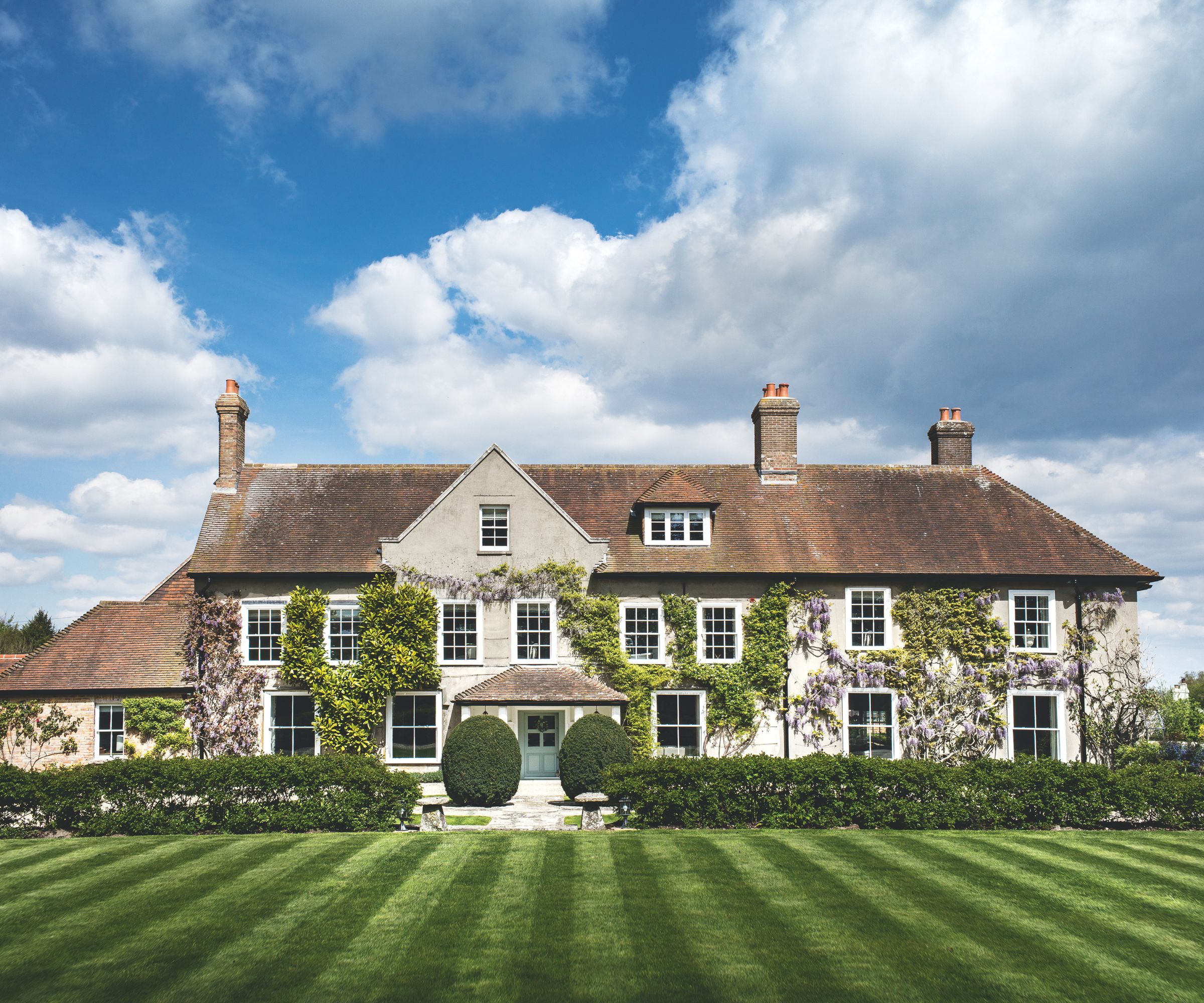 Large country house with wisteria and ivy on the facade, a large freshly mowed lawn, hedges, and a blue door