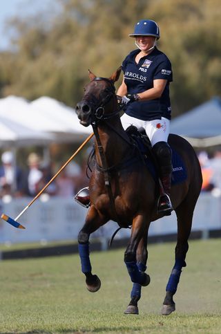 Zara Tindall wearing a blue jersey and helmet while riding a brown horse in a polo match