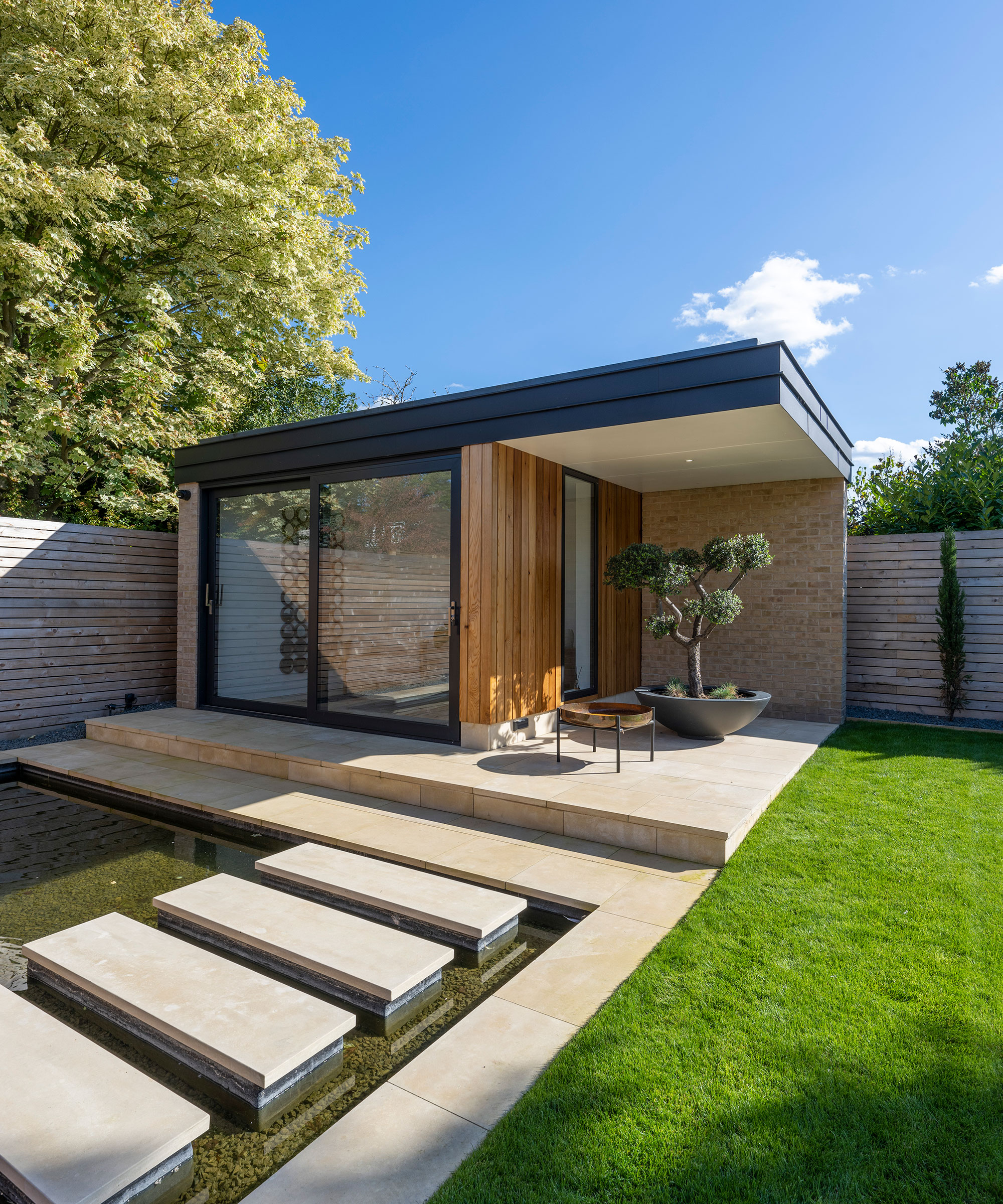 paved stepping stones leading over a contemporary water feature to a bespoke timber-clad garden room with an outdoor firepit