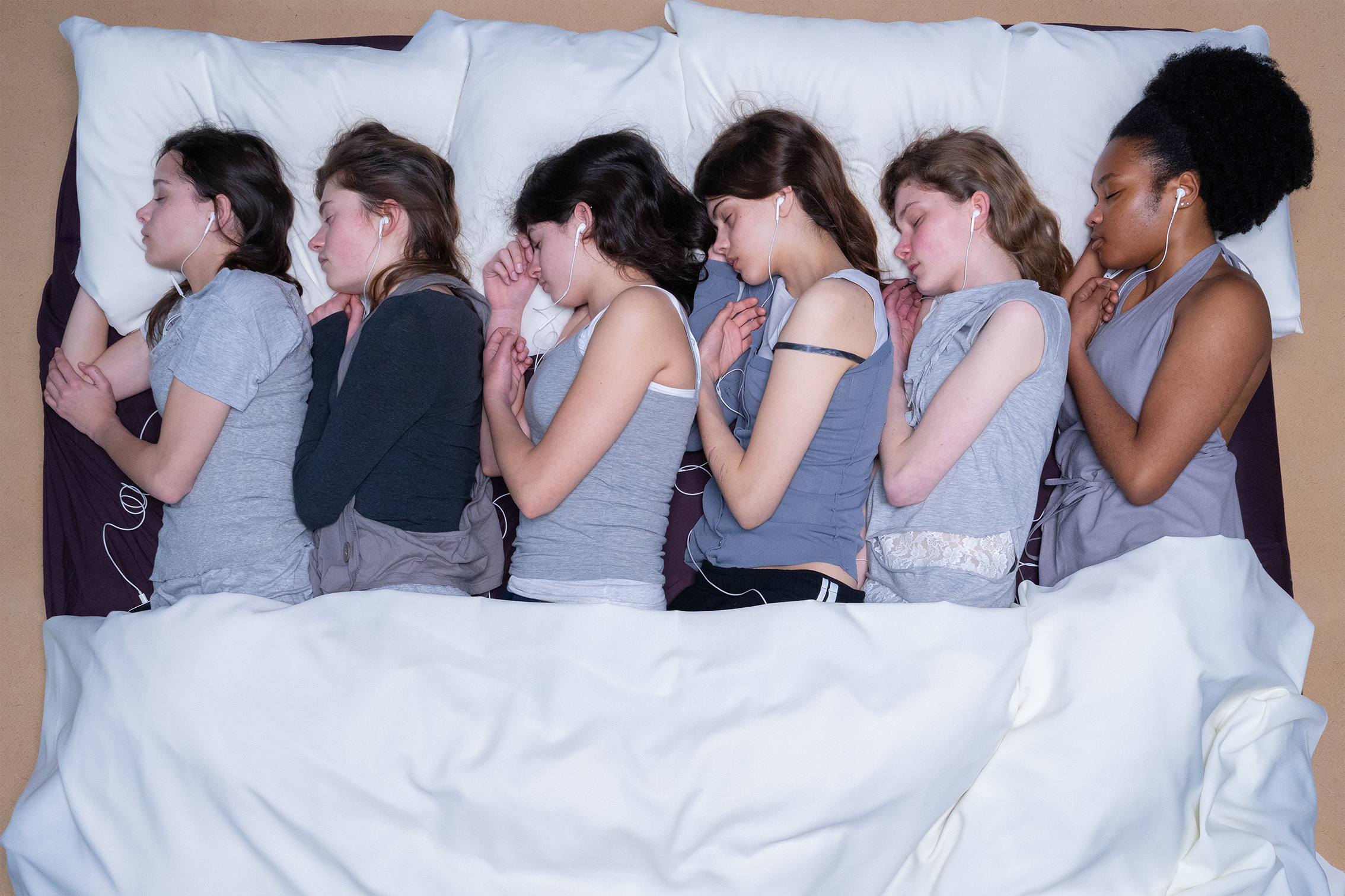 A group of six women in gray tops lie side by side in bed, wearing earphones. Their peaceful expressions and uniform positions suggest relaxation and restfulness