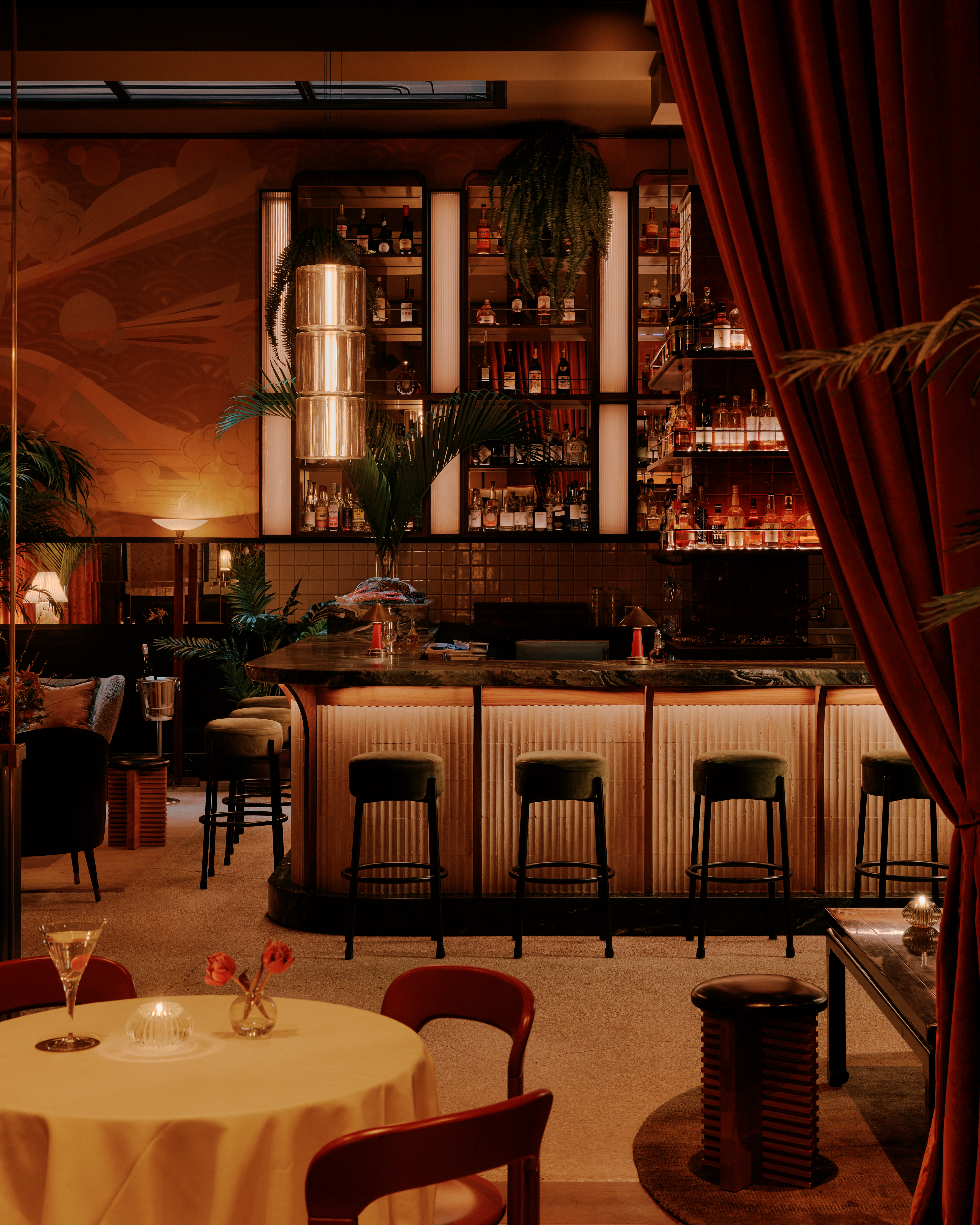 A dimly lit, imaginatively decorated cocktail bar features murals, paintings, Art Deco-inspired lighting, and sprawling plants in a warmly suffused atmosphere.