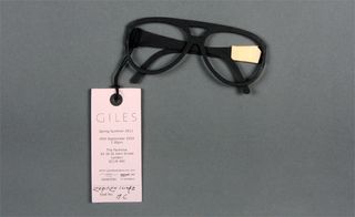 View of the text side of ﻿Giles Deacon's pink label invitation which is attached to a pair of black sunglasses made out of paper. The invitation is pictured against a grey background