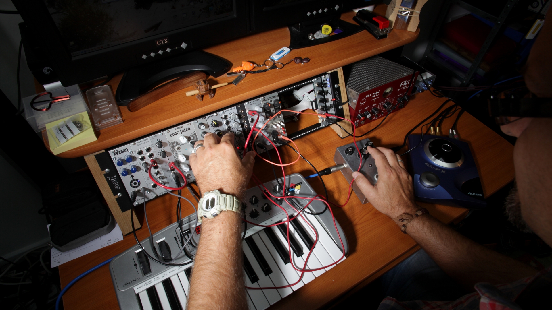 A man sits at a studio desk with various outboard gear, MIDI controllers, and effects