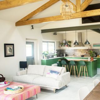 open plan living area with green kitchen, beams, white flooring throughout, pale grey sofa, stripe footstool, dining table to right