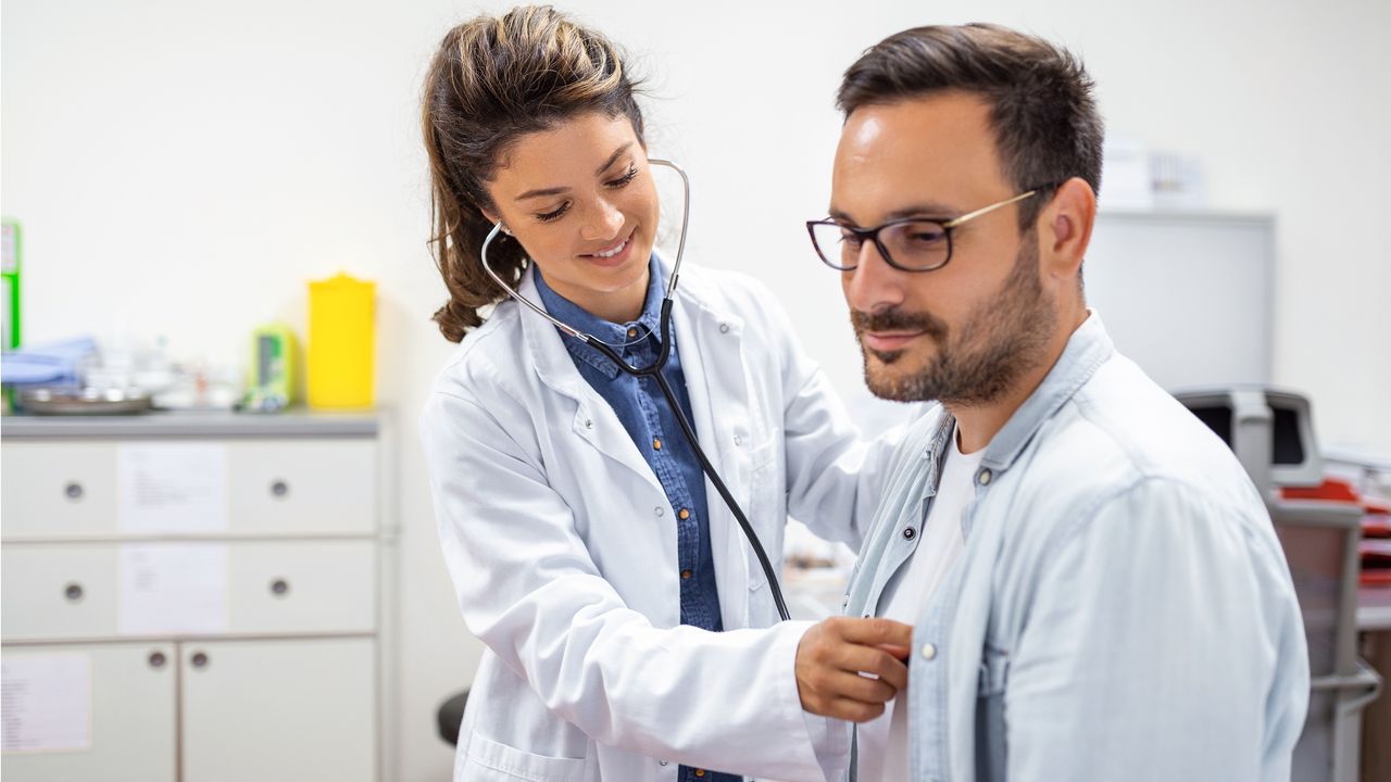 A doctor uses her stethoscope to listen to a man&#039;s heart in a doctor&#039;s office.