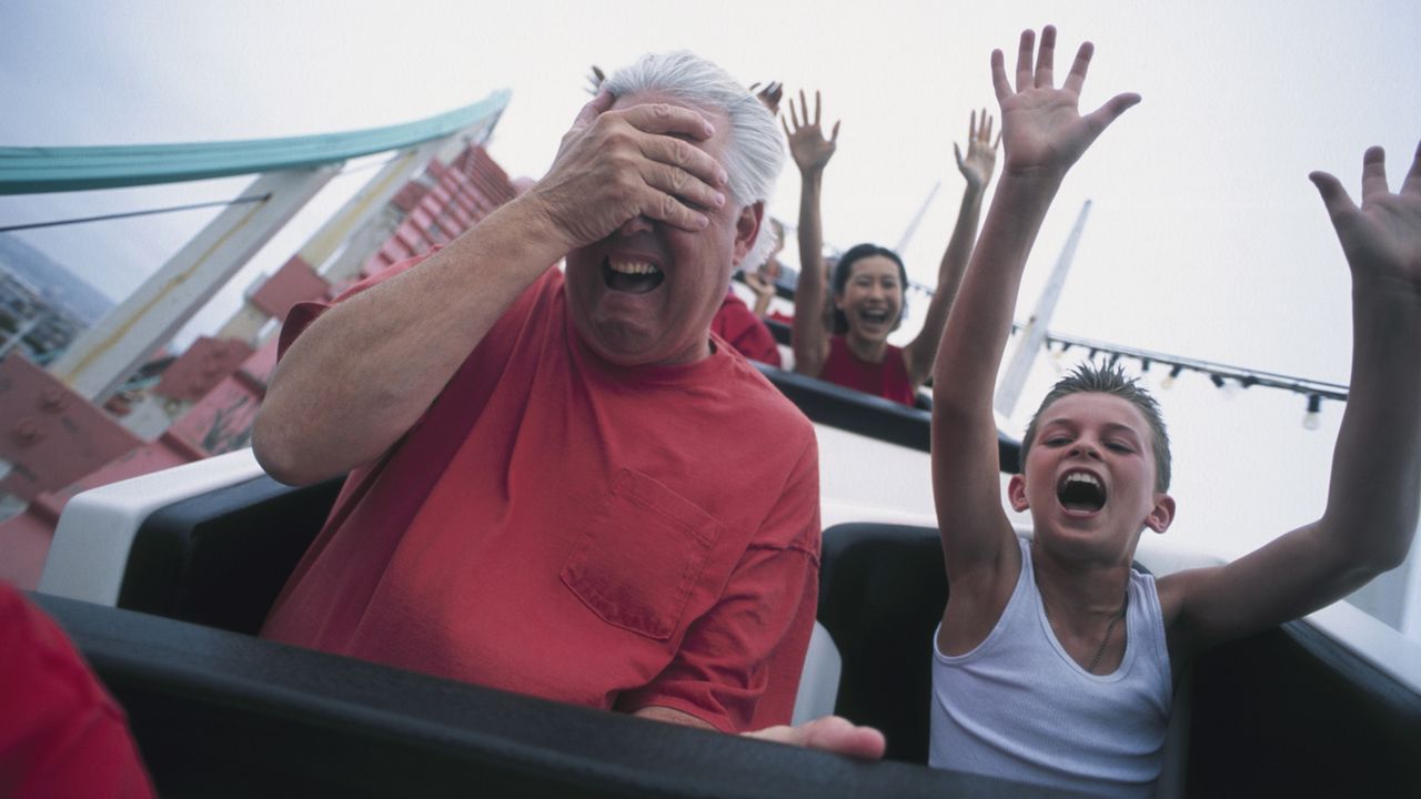 An older man on a roller coaster with a young boy covers his eyes as if he&#039;s scared.