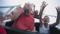 An older man on a roller coaster with a young boy covers his eyes as if he's scared.