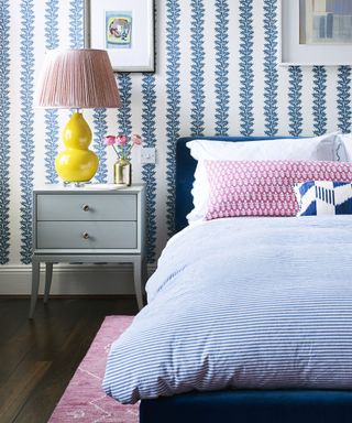 A bed with blue and white striped bedding and and pink pillows next to a grey bedside table with a yellow and pink lamp