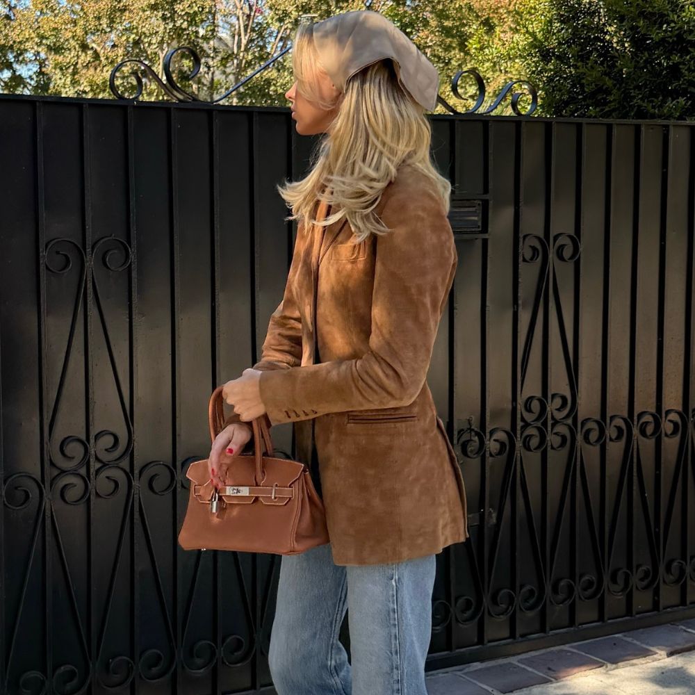 Model Elsa Hosk poses for a photo outside wearing a brown suede jacket with brown loafers, a brown Hermes bag and light-wash blue jeans.