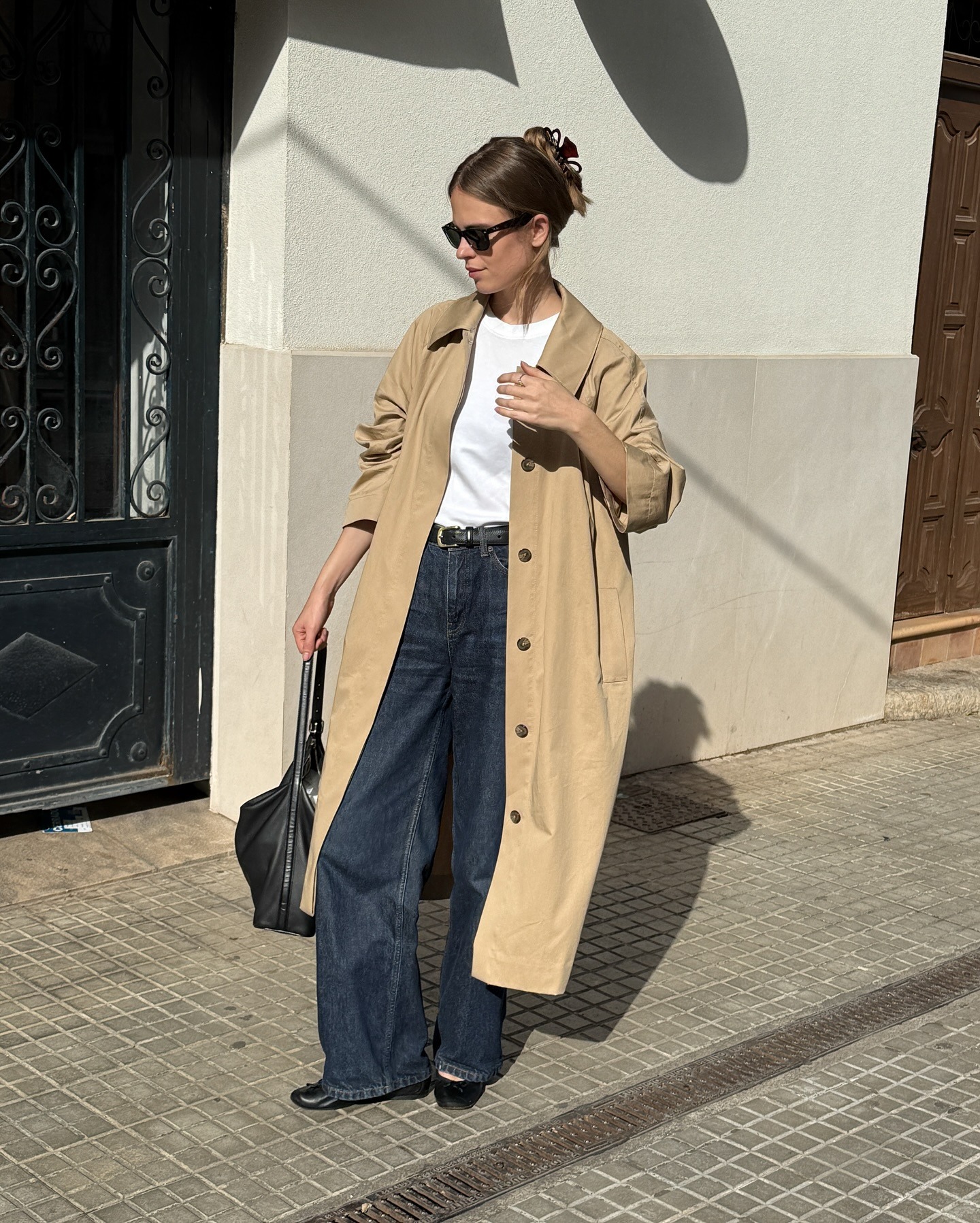 Scandi fashion creative Clara Dyrhauge poses on a sidewalk wearing sunglasses, a trench coat, white T-shirt, black belt, black slouchy shoulder bag, dark-wash jeans, and black ballet flats