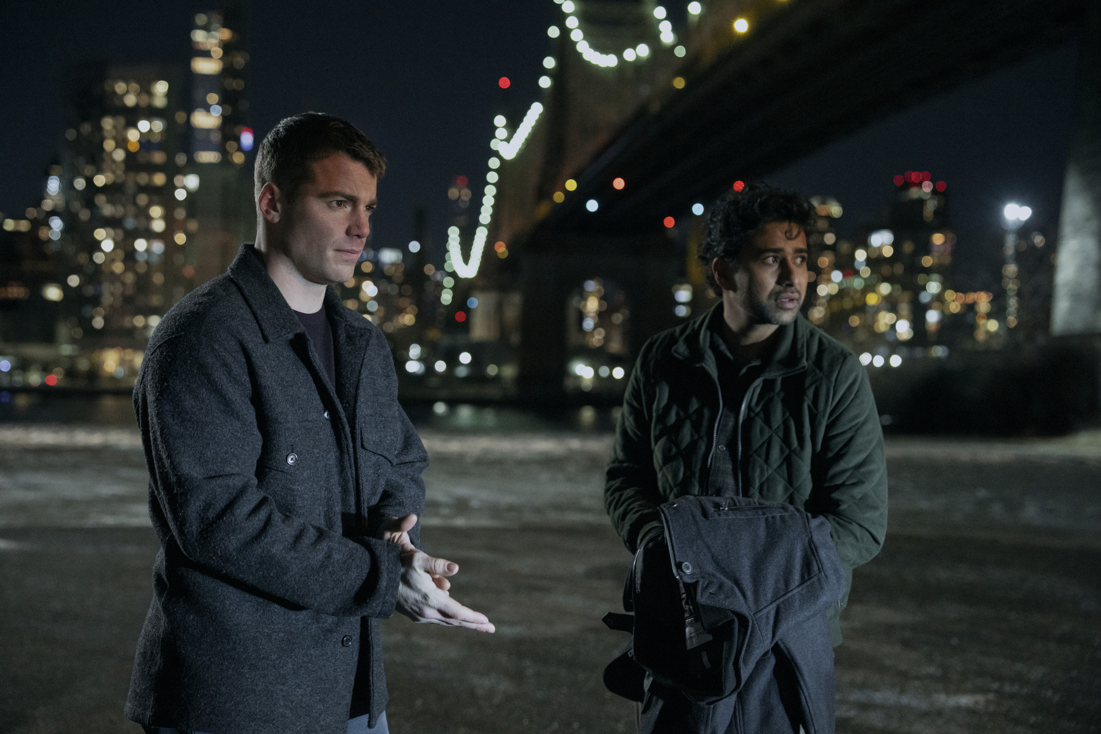 Gabriel Basso as Peter Sutherland and Suraj Sharma as Jay Batra under a bridge with the city lit behind them at night in a still from the night agent season 3
