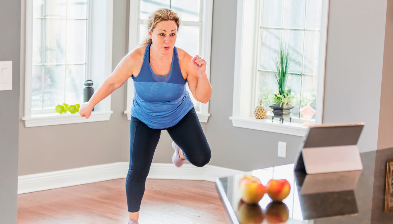 Woman exercising at home in front of a tablet. She is wearing sports leggings and a blue two-tone tank top.