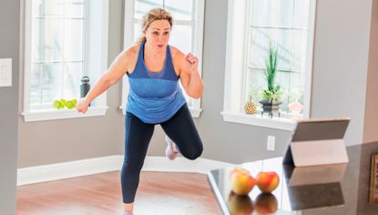 Woman exercising at home in front of a tablet. She is wearing sports leggings and a blue two-tone tank top.