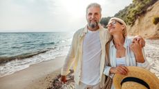 An older couple smile as they walk arm in arm down a beach.