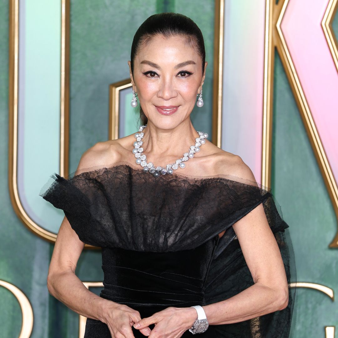 Michelle Yeoh wearing a ponytail hairstyle. She is also wearing a black off the shoulder dress, a diamond necklace, and earrings. 