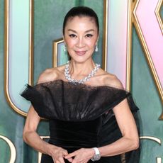 Michelle Yeoh wearing a ponytail hairstyle. She is also wearing a black off the shoulder dress, a diamond necklace, and earrings.