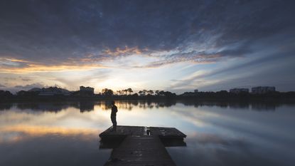 A person stands on a dock watching the sun set over a lake.