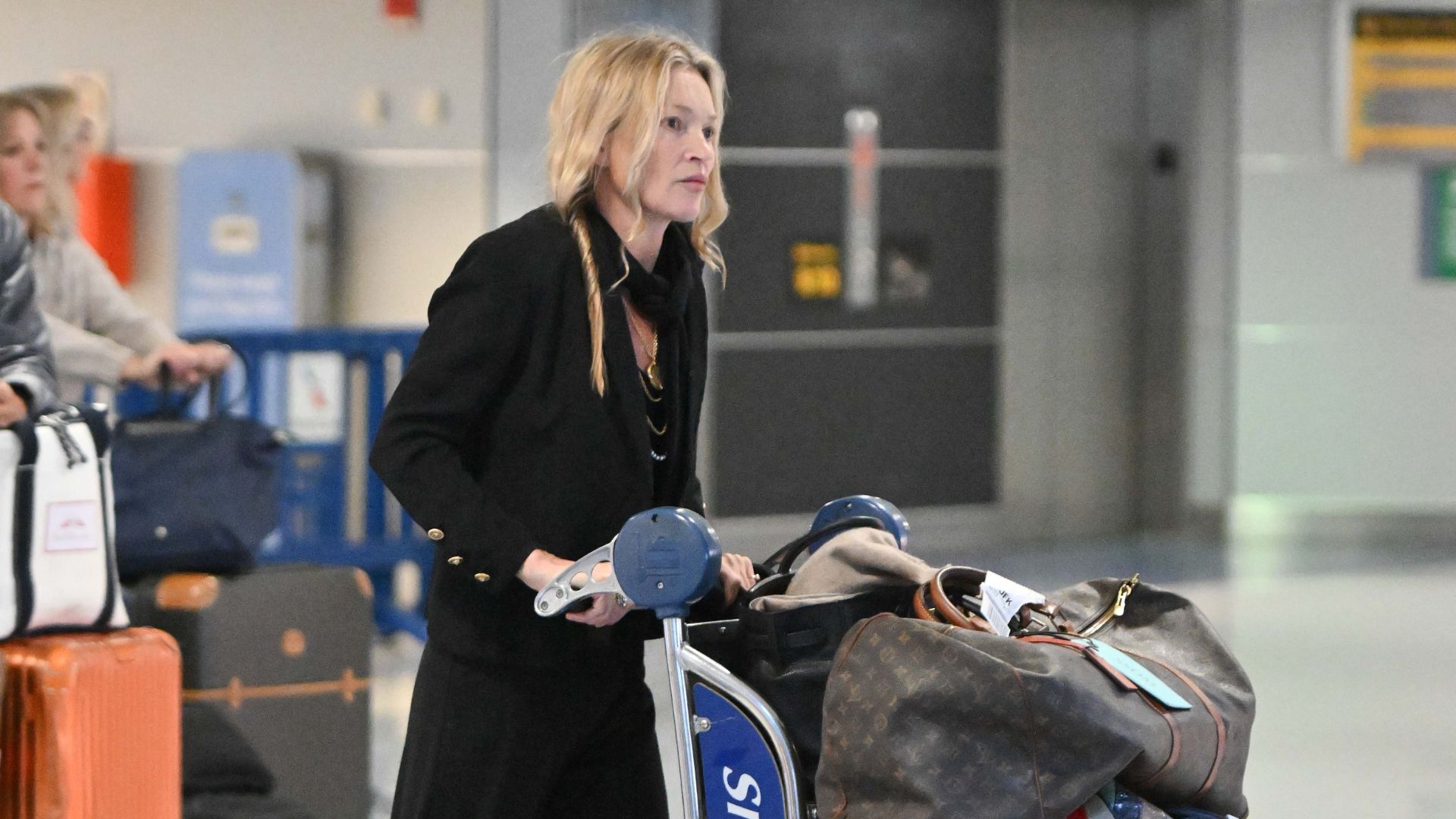 Kate Moss pushes a luggage trolley through the airport wearing black trousers with a black blouse.