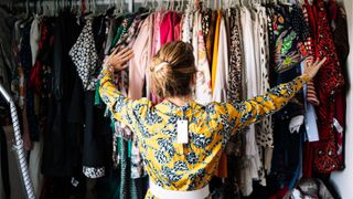 Fashionable blond woman in new yellow dress choosing from clothes rack at apartment -