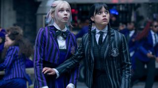 From left to right: Emma Myers as Enid looking up and scared and Jenna Ortega as Wednesday holding her left arm in front of Enid and looking up scared.