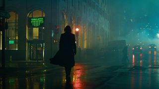 Here we see the silhouette of a man wearing a long trench coat walking on a slick road at night time,