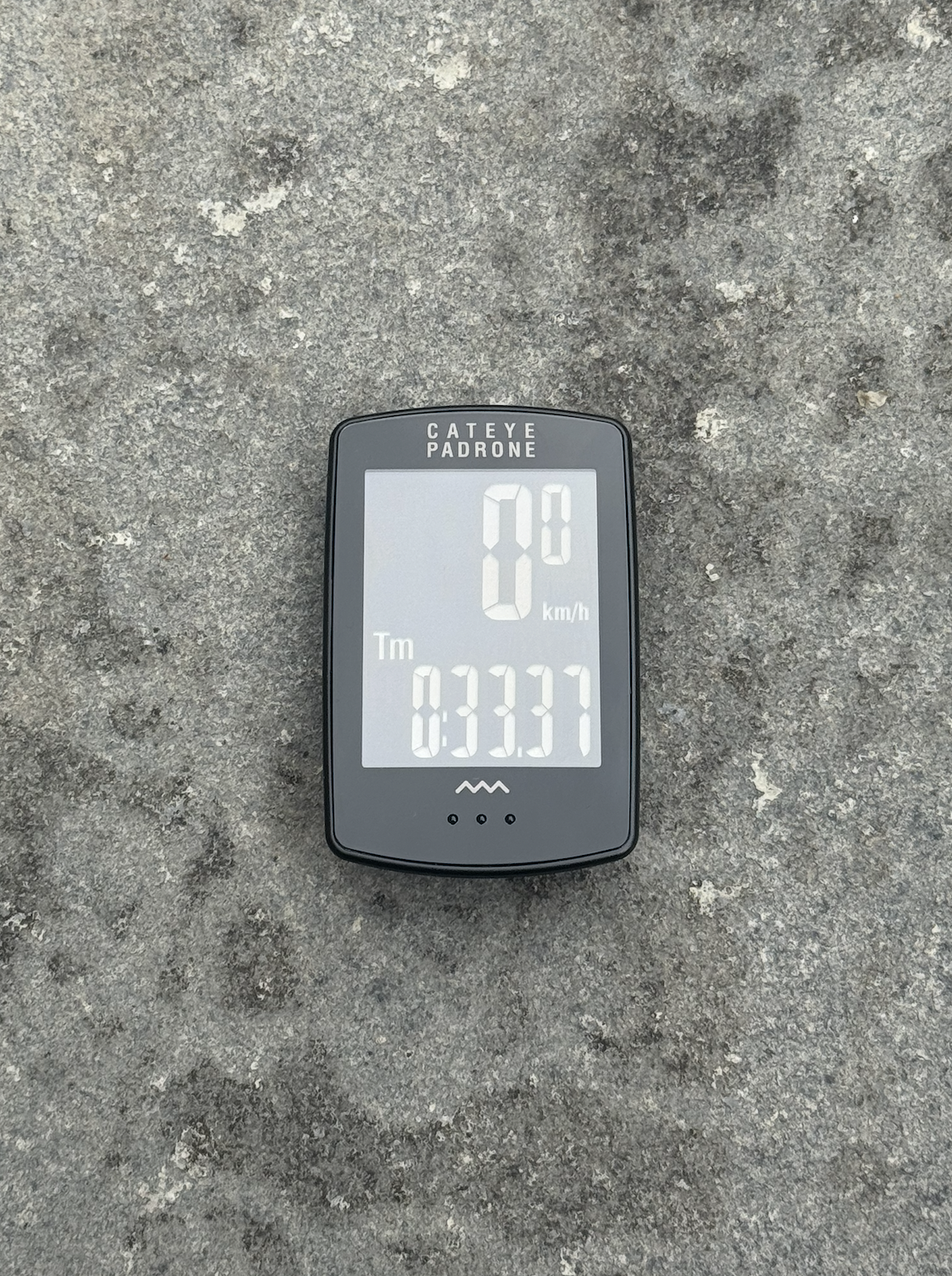 The Cateye Padrone sits on a grey background with the screen lit up and showing km/h and time settings