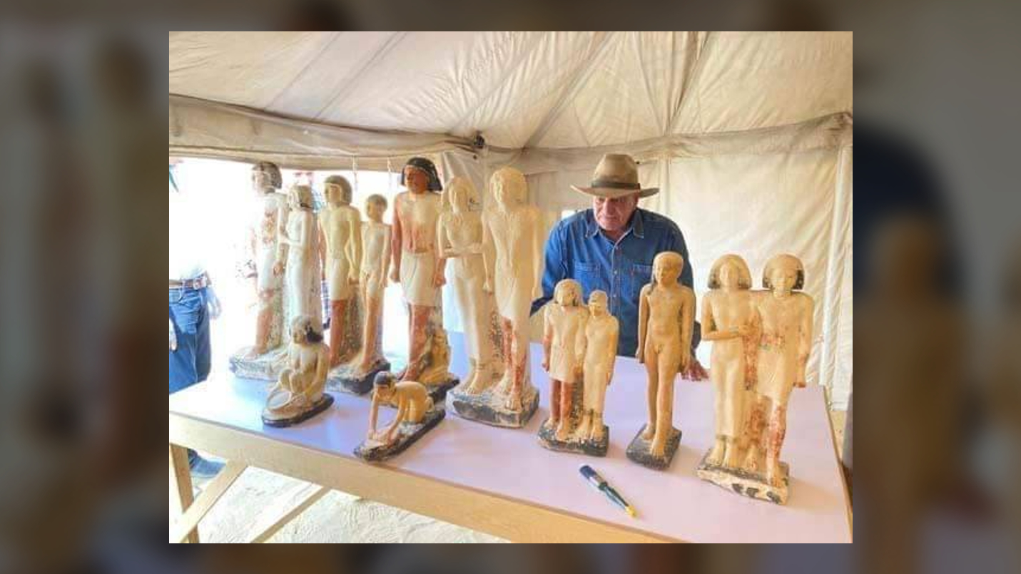 An archaeologist in a blue shirt and hat looks at about a dozen ancient Egyptian statues of people standing on a table.