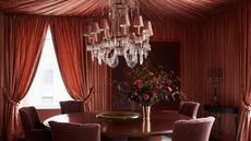 A modern dining room with a tented ceiling in a terracotta color which is continued on to the walls. A large dining table with dark red dining chairs and an opulent chandelier above it.