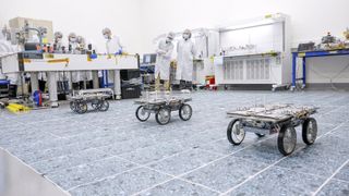 three small four-wheeled rovers sit on a gray floor in a large white-walled room, while people in white lab coats watch in the background
