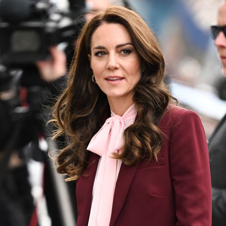 Kate Middleton wearing a maroon suit with a pink tie blouse standing in front of a videographer outside