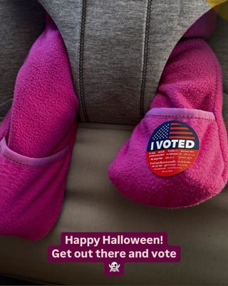 A baby's feet, clothed in a magenta onesie, as he sits in a gray carrier, with an I Voted sticker on one foot.