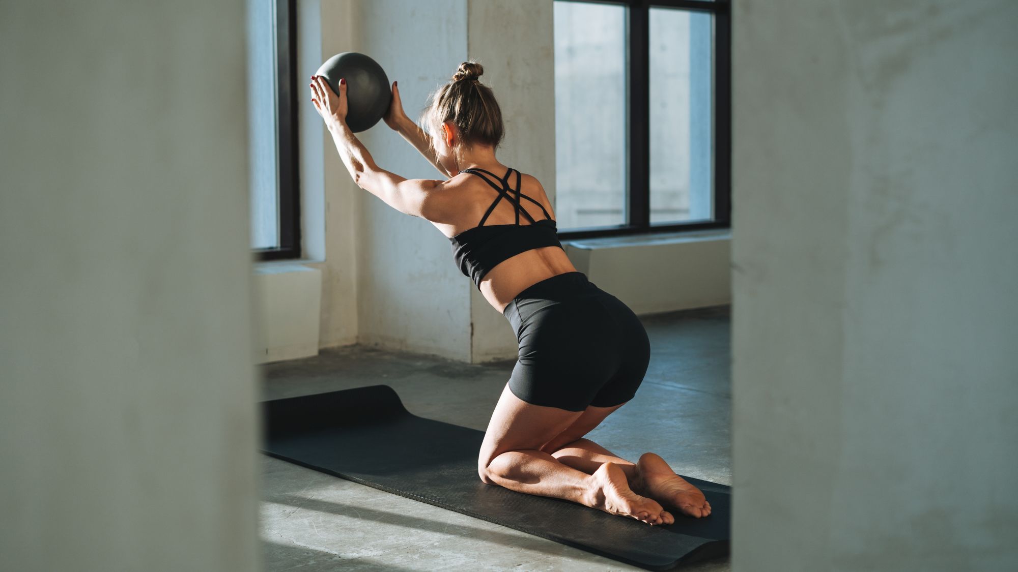 A woman doing some of the best at-home Pilates exercises on a black workout mat with a Pilates ball in hand