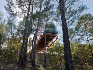 An upward view of a house which is suspended over the edge of a ridge and supported by thick orange metal beams with trees all around it.