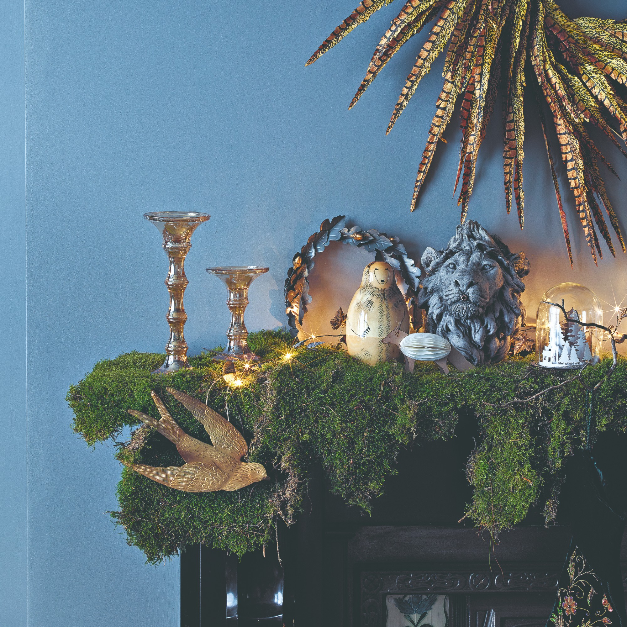 A black fireplace set against a blue wall covered with moss and decorated with Christmas lights, candlesticks and a hammered metal dove decoration