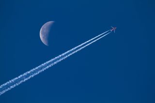 The daytime moon is visible above the contrails of a commercial airplane