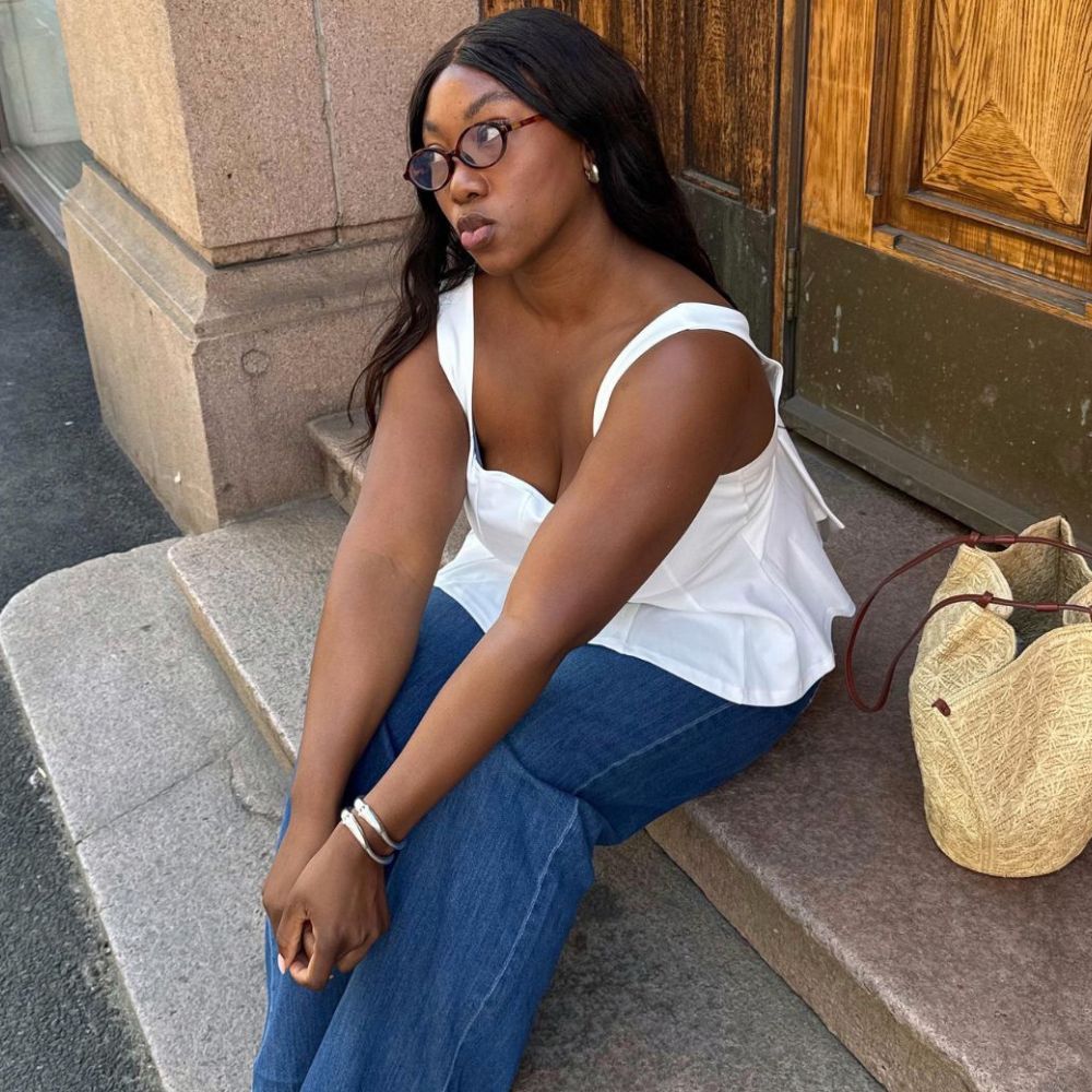 Influencer @nnennaechem sits on steps outside wearing white mesh ballet flats with a white blouse, jeans and a wicker bag.