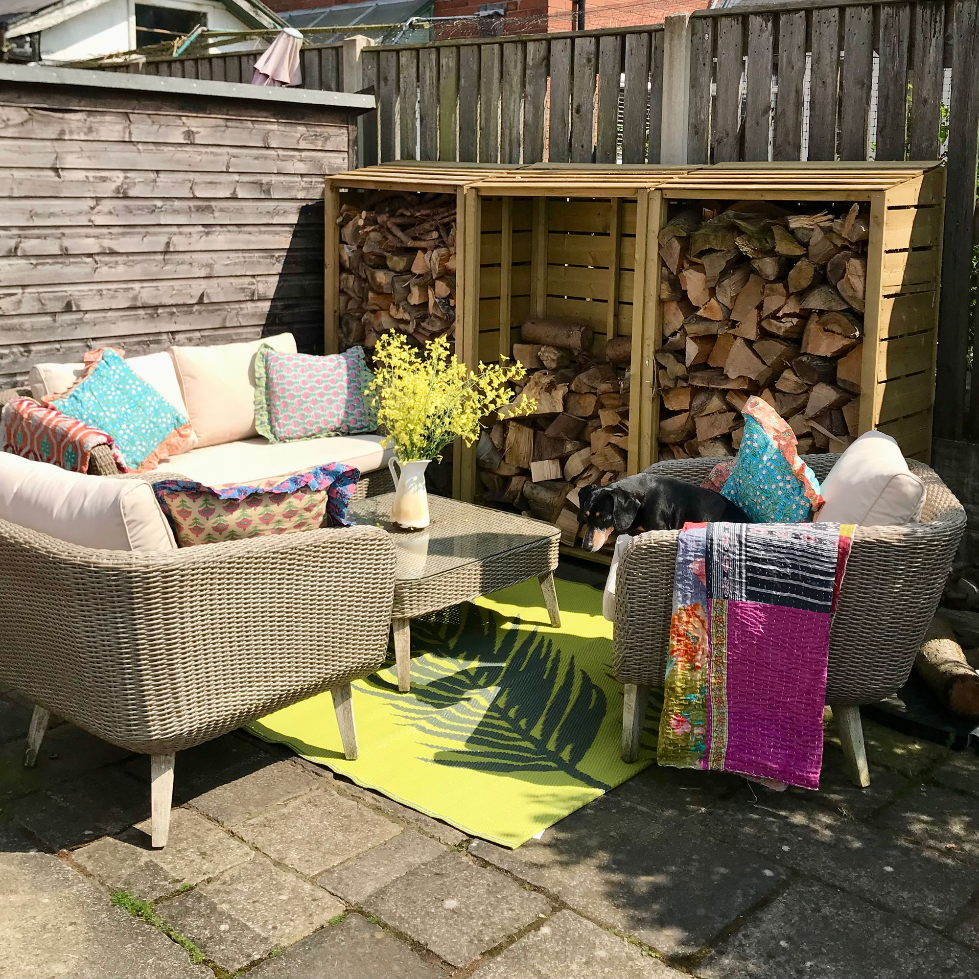 Outdoor sofa and armchairs with colourful cushions and throws around coffee table on top of bright coloured rug