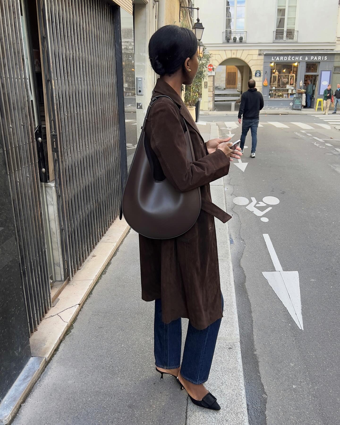 Sylvie wears a brown coat, brown bag, blue jeans, and black kitten heels.