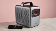 The Anker Nebula Cosmos 4K SE sits on a stone-effect surface with a pink background, with the remote control resting on its side, to the left of the projector.