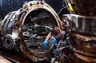 SpaceWorks technician Jerrad Alexander uses a brush to clean an F-1 engine thrust chamber at the Kansas Cosmosphere. Seen in the background, the mist from one of the sprays treating the artifacts with freshwater to remove ocean debris.