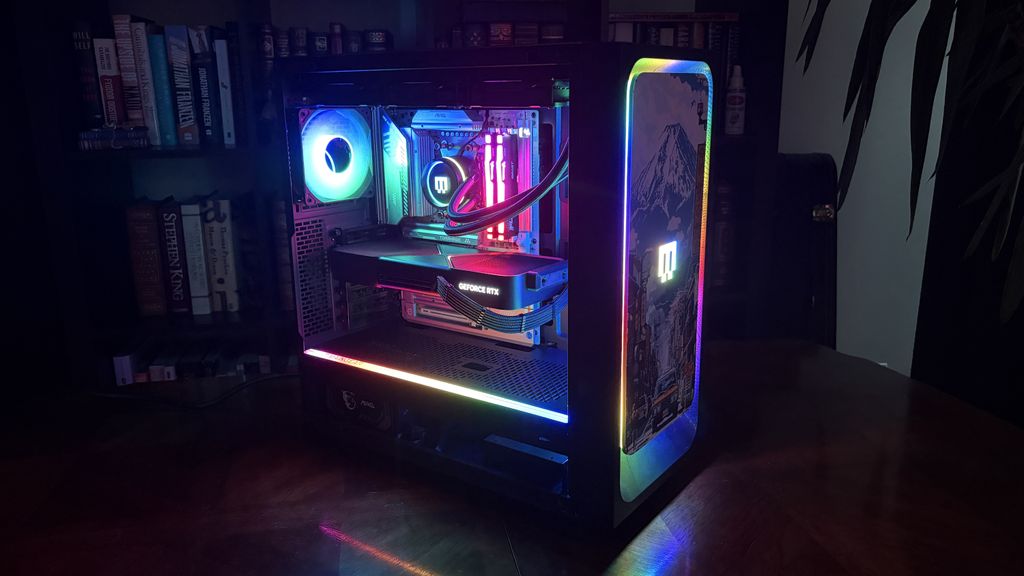 A Maingear MG-1 MK.II gaming PC with colorful RGB lighting inside, illuminating the components in various hues. The setup is on a wooden table, creating a vibrant glow.