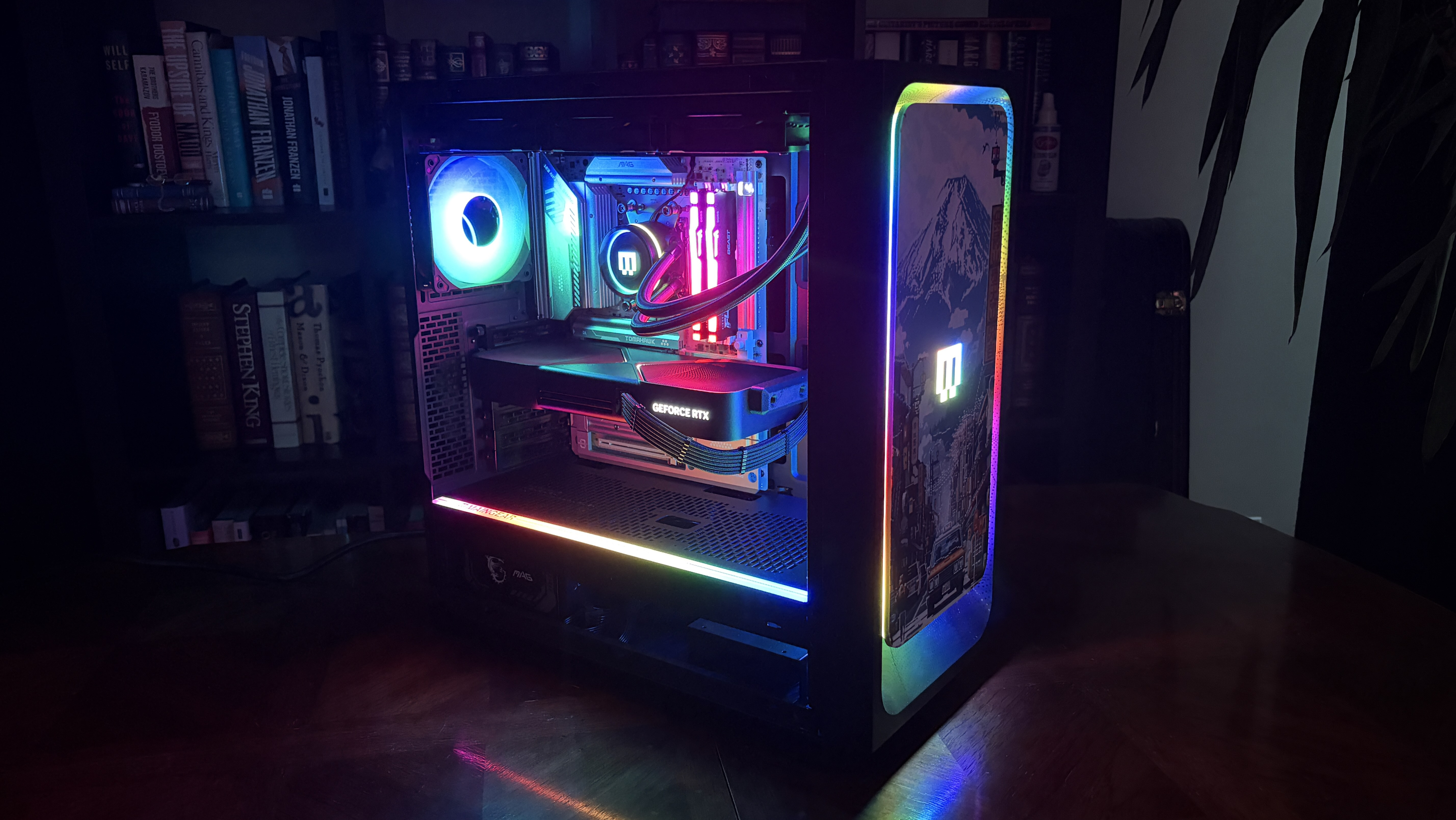 A Maingear MG-1 MK.II gaming PC with colorful RGB lighting inside, illuminating the components in various hues. The setup is on a wooden table, creating a vibrant glow.
