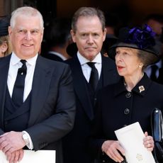 Prince Andrew wears a suit and has gray hair and smirks while his sister Princess Anne wears a black coat and hat and has red lipstick on