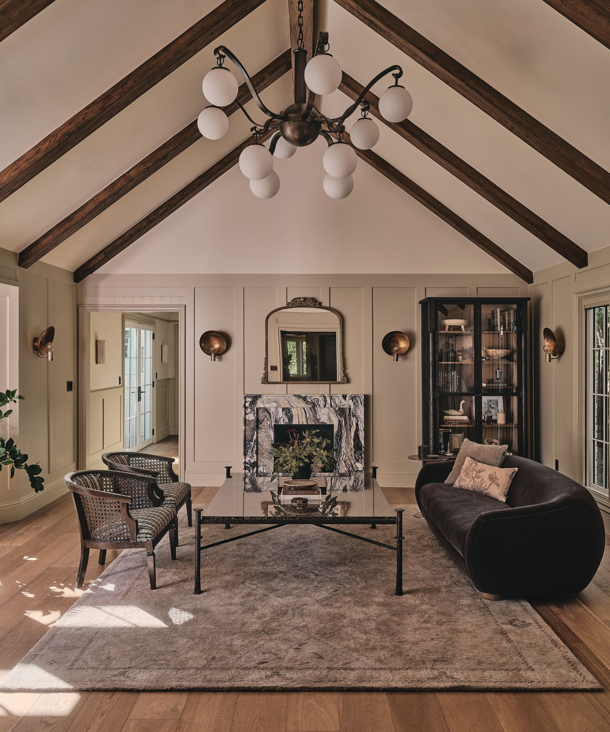 a formal living room with a vaulted ceiling, wooden beams, paneled walls and a veined marble fireplace styled with a curved couch and vintage armchairs