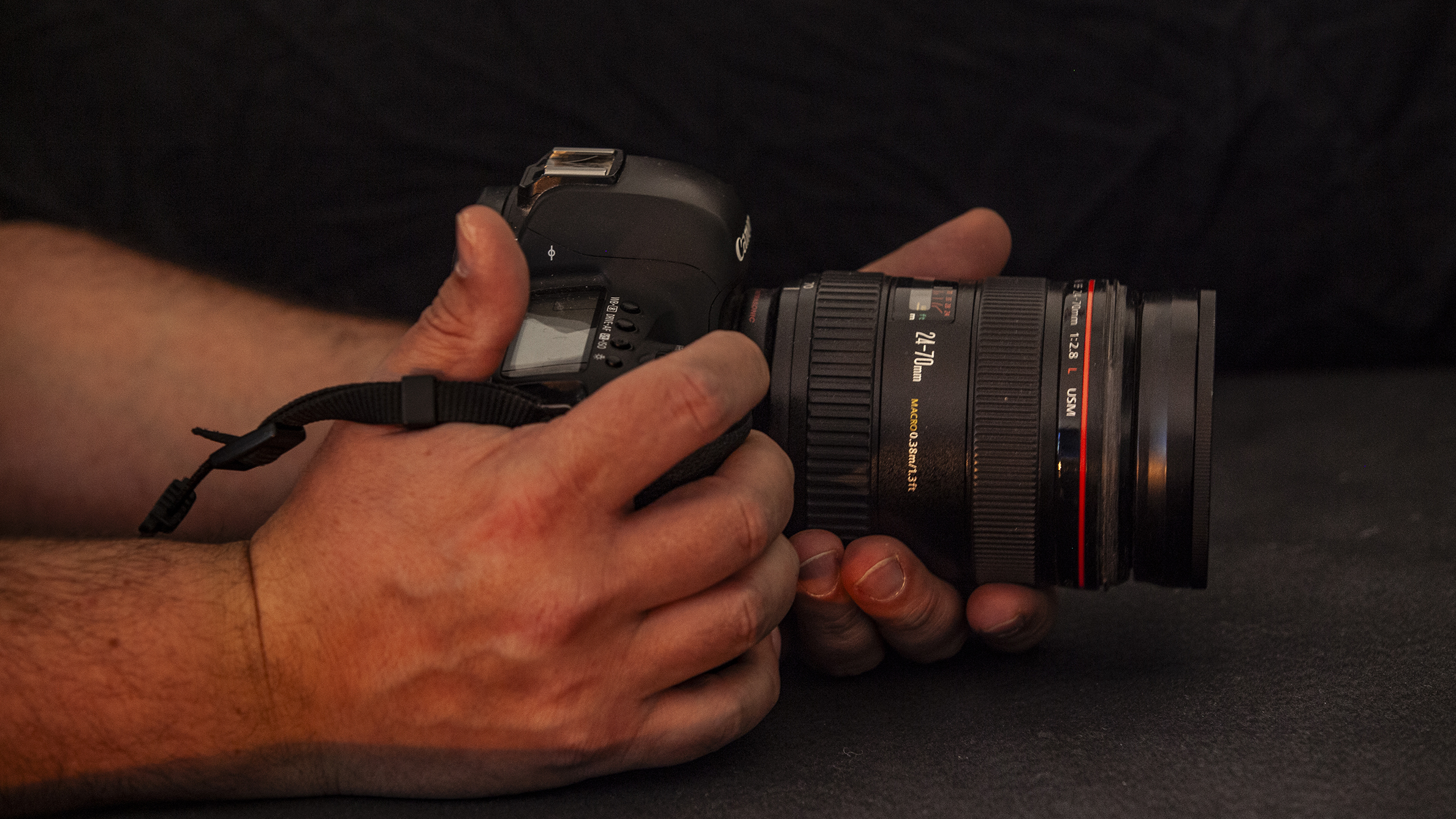 Close-up of hands holding a DSLR camera with a zoom lens, against a dark background. The scene conveys focus and concentration on photography