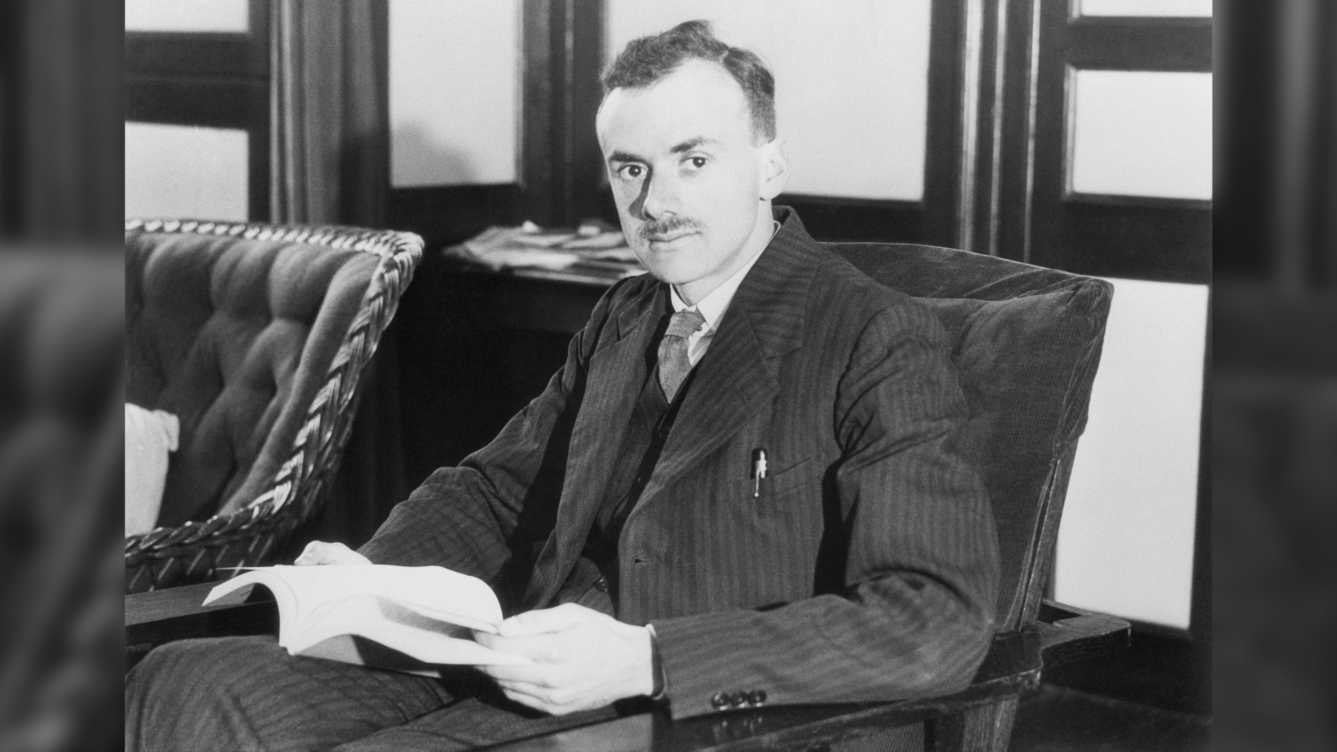 A black and white photograph of Paul Adrien Maurice Dirac. He has short dark hair, a moustache and is wearing a pin-striped suit. He is sitting down in a comfy chair, holding a book open in his lap.