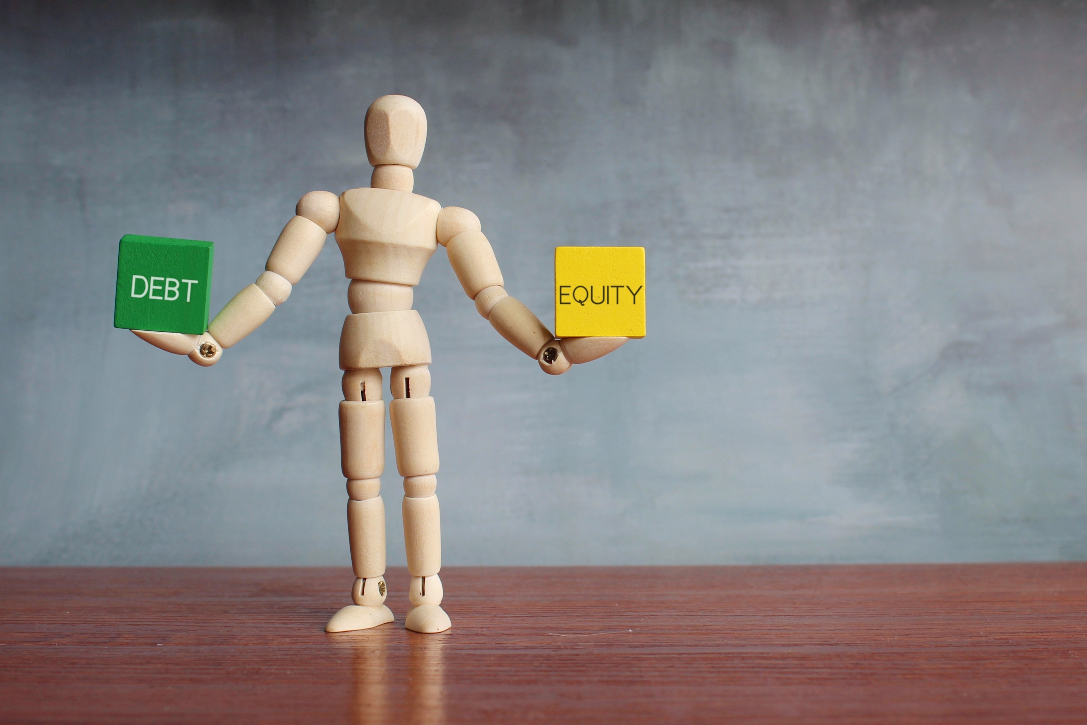 Debt and equity balance concept. Wooden human figure balancing wooden cubes with text DEBT and EQUITY.