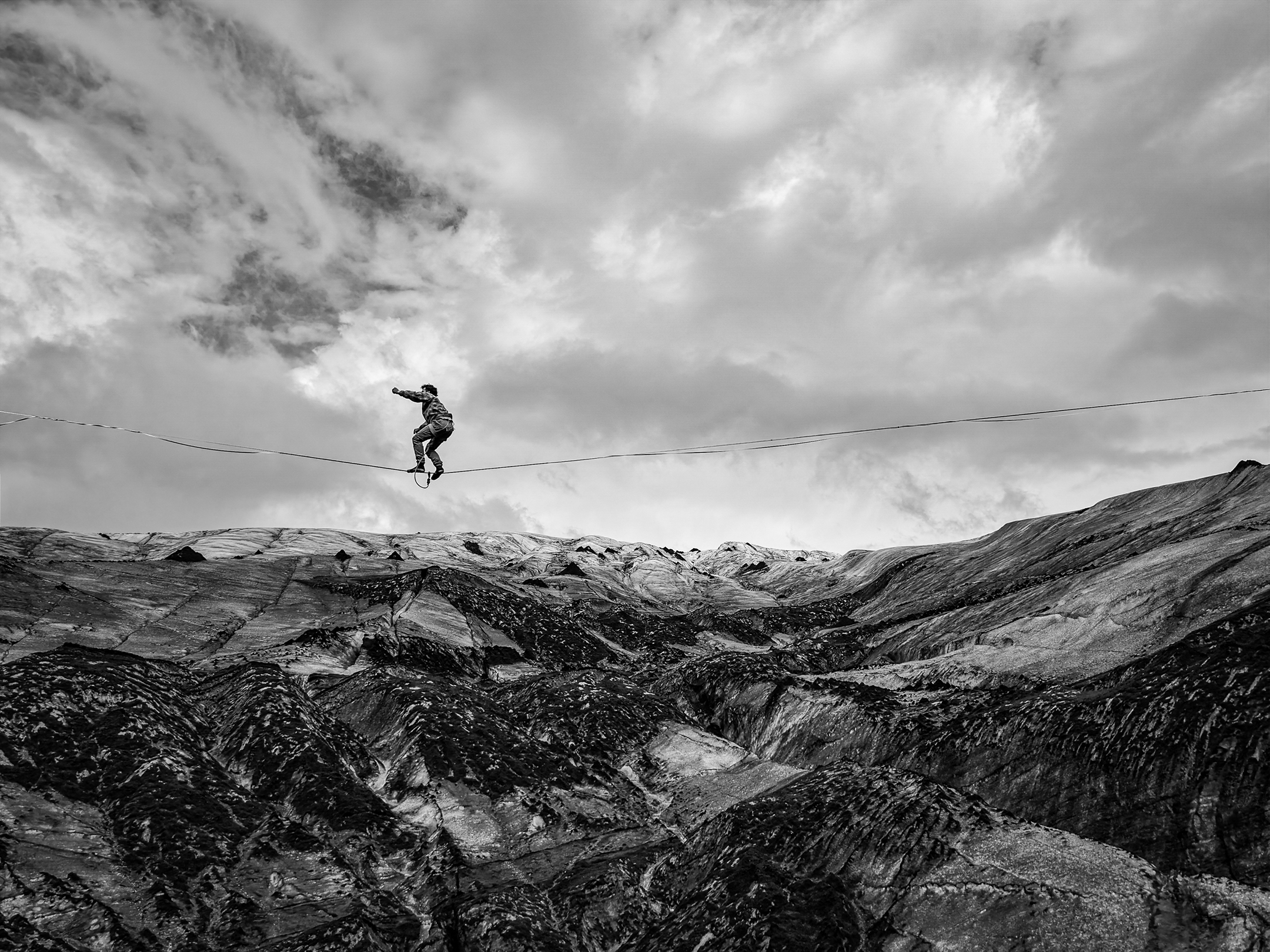 Black-and-white image of a person balancing on a tightrope above a rugged, snow-covered mountain landscape