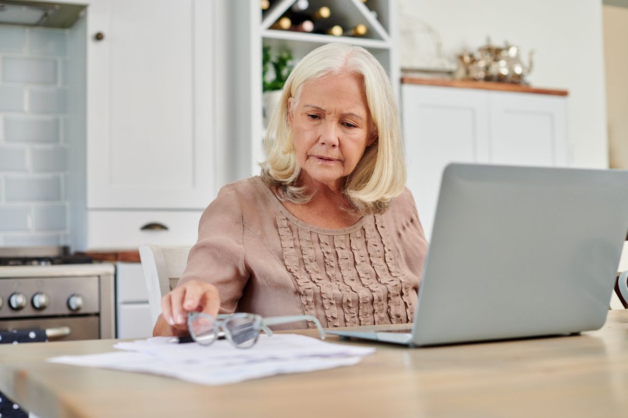 Woman looks worried as she stares at financial document beside her laptop at kitchen table.